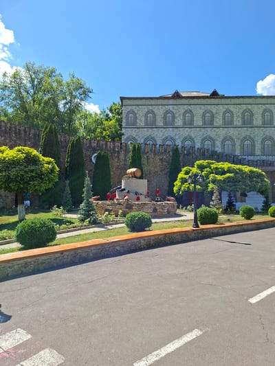 This is a medium shot, eye-level outdoor photograph taken on a sunny day. The scene depicts a courtyard area in front of a building that resembles a castle, located in the city of Milestii Mici, Moldova.

In the foreground, a paved asphalt surface is visible, marked with white lines, suggesting it might be a pedestrian crossing or part of a road. A stone wall, topped with a wooden beam, borders the paved area and separates it from a grassy garden.

The garden features manicured green bushes, small evergreen trees, and a decorative fountain area. The fountain is made of stone and includes a wooden barrel, a wine bottle, and several red wine glasses. Red decorative elements, possibly cannons, are also placed around the fountain.

Behind the garden, a rough stone wall, reminiscent of a castle battlement, stands tall. Beyond this wall, lush green trees form a dense backdrop.

The building in the background is constructed of light-colored stone and has a multi-story facade adorned with numerous arched windows. The roofline is visible at the top, with decorative gables.

The sky above is a clear, bright blue with a few fluffy white clouds. No people are clearly visible, though a single person is partially obscured by a large, round-topped tree on the left side of the frame. The overall impression is one of a well-maintained, themed tourist attraction.