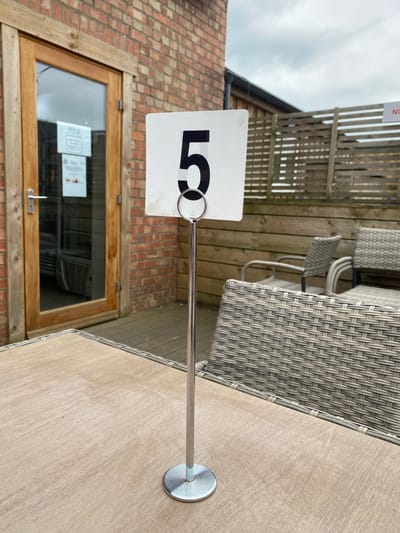 The image shows an outdoor patio area, likely belonging to a restaurant or cafe, in South Oxfordshire, United Kingdom. A table with a woven texture and light wood-grain surface is in the foreground. A table number stand displays the number "5." Behind the table are woven chairs, a wooden pergola, and a brick wall with a wooden-framed window. Two notices are visible inside the window. The sky is overcast.
