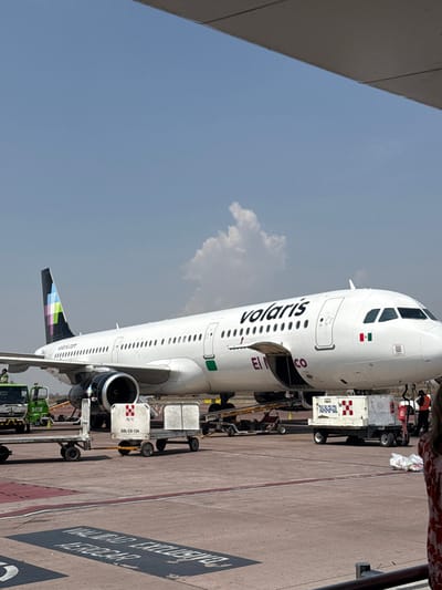 An outdoor daytime scene depicts a white Volaris commercial passenger jet, identified by its fuselage branding and multi-colored tail, parked on an airport tarmac. The aircraft, featuring a Mexican flag decal on its forward fuselage and "El México" text, has both its forward and aft belly cargo doors open. Ground support equipment includes a green service vehicle near the aircraft's tail, a baggage loader conveyor positioned at the forward cargo hold, and multiple white baggage carts, one marked "GOL-CX-124" and another "TAS-09-GP-GDL." Red traffic cones are present on the tarmac. Two individuals in high-visibility vests are partially visible, one near the green vehicle and another near the aircraft's nose, indicating active ground operations. The tarmac is paved, marked with red and white lines, and includes black painted text "VALIDAD EXCLUSIVO AEROCAR..." in the foreground. The sky is clear blue with sparse cumulus clouds. An overhead structure partially obstructs the upper right corner of the frame. A person's back, wearing a white and red patterned garment, is visible in the lower right foreground. The location is an airport tarmac in Tlajomulco de Zúñiga, Mexico, consistent with Guadalajara International Airport.