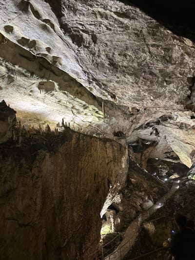 The image depicts the interior of Magura Cave, located near Raianovtsi, Bulgaria. The view is upward, showcasing the cave's walls and ceiling. The cave walls are primarily composed of light-colored rock formations with visible geological features like ridges and depressions.  A constructed walkway is visible near the top of the image, suggesting human modification for tourism or exploration. The lighting is a combination of natural and artificial, casting shadows and highlighting the texture of the rock surfaces. The color scheme is dominated by shades of gray, white, and brown.