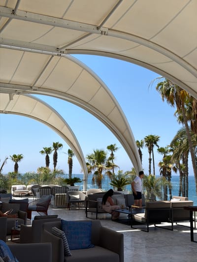 The image depicts an outdoor seating area with a large, white, curved canopy providing shade over a tiled floor. Several people are present, some seated on lounge furniture and others standing. In the foreground, there are comfortable-looking grey armchairs and sofas with decorative cushions. A man in a white t-shirt and dark shorts stands near a seating area, looking out towards the sea. A woman with long dark hair is seated on a sofa with a coffee table in front of her. Palm trees are visible in the background, framing a view of the turquoise ocean and a clear blue sky. The overall setting appears to be a relaxed, possibly resort or beachfront establishment. The location is identified as Pyrgos Community, Cyprus, suggesting a Mediterranean setting.