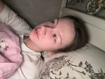 The scene, located in Tlustenkhabl, Russia, depicts a young individual, appearing female, lying on their left side. The person has light skin and light brown hair, and their left eye is open, gazing directly towards the viewer. They are wearing a white t-shirt and a thin, silver-toned chain necklace. Their head rests on a light-colored pillow patterned with dark botanical motifs. A pink blanket or duvet cover, featuring a white cartoon cat head with a pink bow, covers their upper body up to the chest. The background consists of a light-colored headboard or wall panel with ornate, possibly gilded, decorative elements, indicating an indoor setting, likely a bedroom. The lighting is soft and ambient.