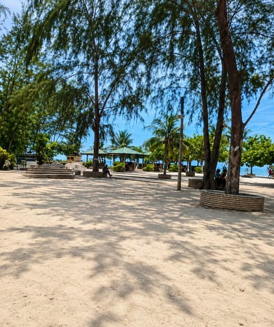 The image depicts a sandy area with scattered trees and palm trees, suggesting a tropical beach or park setting. In the background, there are several open-air pavilions with green roofs, some of which appear to be seating areas. A few individuals are visible: one person is sitting on a stone bench near a tree, another group of people are gathered near a tree, and a couple of individuals are walking in the distance. The ground is covered in sand, with shadows cast by the trees. A concrete structure, possibly a planter or seating area, is visible in the foreground. The sky is blue with some visible clouds. The overall scene suggests a public outdoor recreational space. The location is identified as Kota Pari, Indonesia.