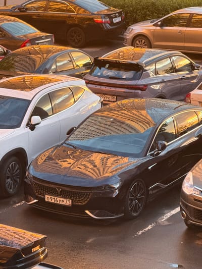 This media file depicts a vertical, eye-level shot of several parked cars on a paved surface, likely a street or parking lot, in Drozhzhino, Russia. The scene is bathed in the warm, golden light of either sunrise or sunset, casting long shadows and creating strong reflections on the cars' surfaces. The sky appears clear, with no visible clouds.
The primary subjects are the cars themselves. A silver SUV is parked on the left, partially obscured by grass. Next to it are several darker-colored cars, including a black sedan with a prominent front grille and a license plate reading "0916 YP 71 RUS". The reflections on the cars show the facade of a multi-story building, suggesting an urban or suburban environment. Another white vehicle is parked to the right, also reflecting the building. A dark-colored car with a "THULE" logo visible on its rear is also present.
The activity taking place is simple: the cars are parked. There is no human presence or overt action. The notable details include the strong directional light, the reflections on the cars, and the Russian license plate, which provides a specific location clue. The overall mood is tranquil, perhaps even serene, due to the peaceful setting and the quality of the light. The image captures a moment of stillness in an everyday urban environment.