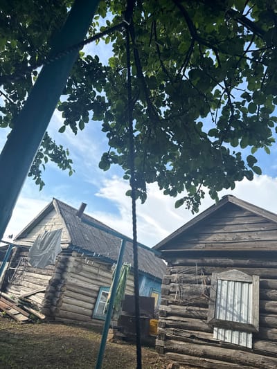 In Чуй-Атасево, Russia, an outdoor scene depicts two rustic wooden log houses under a bright blue sky with scattered clouds. Lush green tree foliage partially obscures the view from above, with branches and leaves dominating the upper portion of the image. A thick, dark rope hangs vertically in the foreground, likely part of a swing set or similar structure, casting a shadow on the ground.
The house on the left, with a gray, weathered roof, has laundry drying on a clothesline in front of it, including a large white sheet. A small blue-painted structure is visible behind it, and the log construction of the house is prominent, showing the rough-hewn texture of the timber. A narrow, blue-framed window can be seen.
The house on the right, also constructed of dark, aged logs, features a wooden-framed window with vertical, corrugated metal slats. Above the window, a rusty metal rack is affixed to the wall. The ground in front of both houses appears to be dirt and dry grass. The overall impression is one of a rural, possibly dated, residential area.