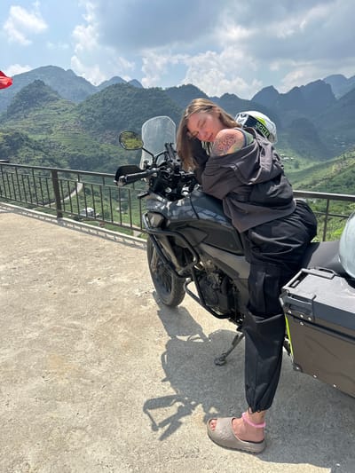 A woman with brown hair is resting on a motorcycle in Lũng Phìn Commune, Vietnam. She is wearing a grey jacket, black pants, and grey slides. Her head rests on the gas tank and she has her helmet behind her head. Her left arm, featuring a tattoo, rests on the motorcycle. The motorcycle is black and has an aftermarket cargo box attached to the rear of the seat. A black railing separates the foreground from a mountainous backdrop.
