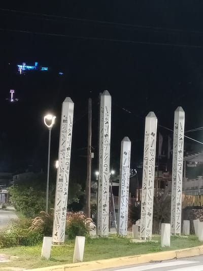 At night, the image captures a set of five tall, white rectangular posts, each resembling a giant pencil, adorned with drawings of school supplies and letters. These are located on the median of a street in Villa Carlos Paz, Argentina. The posts decrease in height from back to front. A street light is positioned in front of the nearest post. The posts are surrounded by grass. In the background, there is a dark sky, utility lines, and street lights.