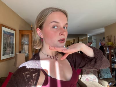 A fair-skinned person with blonde hair and blue eyes is positioned upside down in the foreground. She is wearing a burgundy tank top and a brown crocheted sweater. She has a red and silver necklace and a silver ring on her left hand, which is raised towards her face. Her ear is adorned with multiple earrings. The background includes a living room setting with a couch, a glass cabinet, a wooden wall clock, and framed artwork. The scene appears to be indoors during daylight hours. No specific indicators confirm this is in Kelowna, Canada.