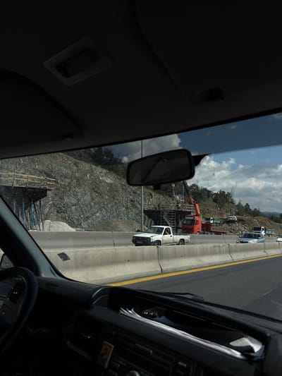 The image captures a bright daylight scene from the interior of a vehicle, looking out through the front windshield. The view is oriented such that a multi-lane highway runs along the right side, separated by a concrete barrier with a prominent yellow line. Various vehicles are visible on the highway, including a white pickup truck in the foreground right with the partial license plate "TAO 3098", a silver car, and several other trucks or construction vehicles further down the road.

To the left of the vehicle, a large, steep, rocky hillside dominates the midground. Extensive construction activity is underway on this hillside, featuring scaffolding structures, an orange crane truck, and other earth-moving equipment. Numerous white rectangular markers are visible on the rock face, possibly indicating drilling points or geological survey locations for stabilization or road expansion. Trees are scattered along the top of the hill.

The sky above is bright blue with scattered white clouds, suggesting clear and sunny weather. Inside the viewing vehicle, parts of the dashboard, the rearview mirror, and the headliner are visible, framing the external scene. The setting appears to be a major road or highway traversing a mountainous or hilly region, undergoing significant infrastructure development or maintenance in the Agios Pavlos Municipal Unit, Greece. No specific individuals are clearly discernible, but the presence of heavy machinery implies ongoing work.