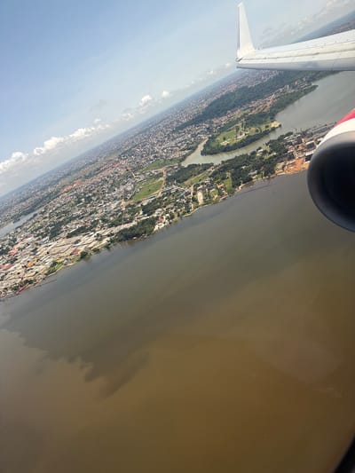 The image shows an aerial view of Abidjan, Ivory Coast, taken from an airplane. Visible are a city shoreline, a large body of water, and parts of the aircraft, including a wing and engine. The city has dense buildings and green spaces. The water has a murky appearance.