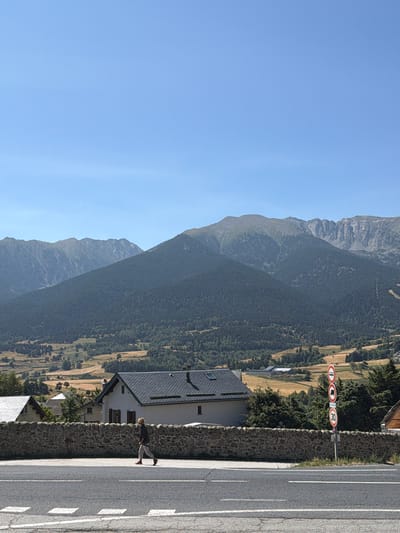 In Mont-Louis, France, a person walks along a paved road on a clear, sunny day. The road has white lane markings. Behind the road, a stone wall separates it from a residential area. In the background, there are houses with dark tiled roofs, and further back, a lush green forest covers a large hill. Beyond the hill, imposing mountains with rocky peaks rise against the blue sky. Several traffic signs, including a "30" speed limit sign, are visible on the right side of the road. The person is wearing a dark long-sleeved shirt and light-colored pants, with red shoes.