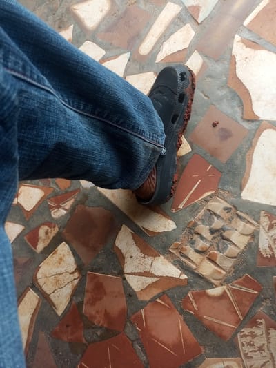 A low-angle, close-up shot shows a person's left leg clad in blue denim jeans and a black clog shoe, partially covered in reddish-brown dirt, resting on a floor composed of irregularly shaped, broken terracotta tiles and grey cement. The tiles display variations in color, ranging from pale beige to deep red, with some exhibiting cracked surfaces and chipped edges. The overall impression is of an outdoor or semi-outdoor setting, possibly a patio or walkway in Agbara, Nigeria, where the dry, dusty conditions are evident from the dirt on the shoe and floor.