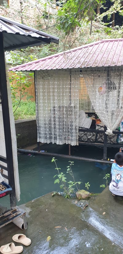 In a shaded outdoor setting, likely a cafe or seating area near water, a young person wearing a blue and white shirt with cartoon graphics is crouched down, holding a phone and appearing to take a photograph or video. The person is positioned on a wet, concrete surface adjacent to a dark, still body of water. This water contains some visible debris, including bottles and other refuse floating on the surface and along the edge.
Overlooking the water is a covered seating area with a red, corrugated metal roof. The structure is partially enclosed by white lace curtains, and inside, cushions and what appears to be seating are visible. To the left, a dark metal frame supports another section of roofing and a white cloth.
The environment is lush with greenery, including trees and plants growing along the water's edge and behind the structures. The background features a brick wall and more foliage, suggesting an urban or semi-urban location with natural elements. The overall scene conveys a relaxed, possibly slightly unkempt, outdoor atmosphere. The location is identified as the city of Avval', Uzbekistan.