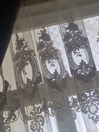 The image shows a close-up of a sheer lace curtain with an ornate floral pattern. The curtain is grey and white, and it is hanging in front of a window. The pattern features large roses and oval frames with intricate lace detailing. The background appears to be a window with blinds, allowing light to filter through the translucent fabric. The scene is indoors. The location in Narchak, Uzbekistan is not discernible from the image.