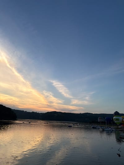 This is a medium shot, captured from a low angle, looking across a body of water towards a distant shoreline. The sky is a clear blue with wispy white clouds, particularly prominent on the left side of the frame where they are illuminated by the setting sun with hues of orange and yellow. The water's surface is choppy, reflecting the sky and the dark, tree-covered hills that form the shoreline in the middle ground. Several boats are visible on the water, some moving and leaving wakes, while others appear to be docked. Along the shoreline, a cluster of colorful hot air balloons are the most prominent objects in the scene. One balloon, predominantly green and white with Polish text and a logo, is particularly detailed. The environment appears to be a lake or large river in a rural or semi-rural setting, identified as Gródek nad Dunajcem, Poland, likely during a hot air balloon festival. No people are clearly discernible as individuals, but small groups may be present near the balloons and boats. The overall impression is a serene twilight scene with a touch of festive activity.