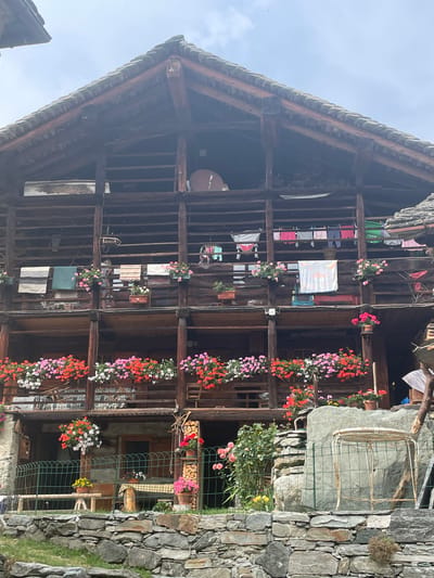 The image depicts the exterior of a traditional wooden chalet in Alagna Valsesia, Italy, against a backdrop of a cloudy sky. The chalet features multiple levels of wooden balconies adorned with numerous potted plants and hanging flower baskets. On the balconies, various items are hung, including laundry and what appear to be textiles or rugs. A person is partially visible on one of the upper balconies. The foreground is characterized by a stone wall, grass, and a green wire fence enclosing a small area with a metal table and chairs. The architectural style suggests a rustic, possibly historical, mountain dwelling.