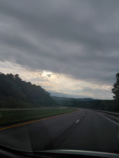 An eye-level, wide-angle shot captures a highway winding through a forested, hilly landscape under a cloudy sky. The road, paved with asphalt, is dark and wet, suggesting recent rain. A solid yellow line demarcates the left edge of the travel lanes, while dashed white lines mark lane dividers. A metal guardrail runs along the right side of the road. Lush green trees and vegetation cover the slopes on the left, with more distant, rolling hills visible in the background. The sky is predominantly overcast with dark, heavy clouds, but a patch of brighter sky, possibly with sunlight breaking through, is visible in the upper left quadrant. A single white vehicle is seen in the distance on the left side of the highway. The photograph appears to have been taken from inside a vehicle, with the lower portion of a windshield and dashboard visible in the foreground. The scene is devoid of people.