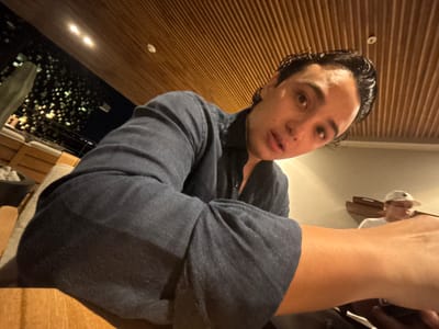A close-up, low-angle selfie features a man with dark hair and olive skin wearing a dark gray shirt. He is looking at the camera. He is at a wooden table or bar, possibly in Cabo San Lucas, Mexico. Outside, lights from buildings shine from what appears to be the city at night. In the upper left corner of the photo are the arm and hand of another person.