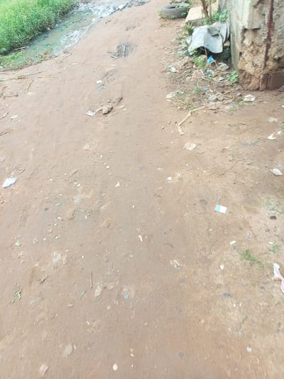 This is a ground-level, eye-level shot of a dirt road in Agbara, Nigeria, on what appears to be a cloudy day. The road is unpaved and uneven, with a ditch filled with murky water and grass on the left side of the frame. Scattered on the road are various pieces of debris, including plastic wrappers, what look like discarded food packaging, and some larger pieces of scrap metal or discarded items near a stone wall on the right. The wall itself appears weathered and shows signs of rust. There are no people visible in the scene. The overall impression is one of an undeveloped or neglected area.
