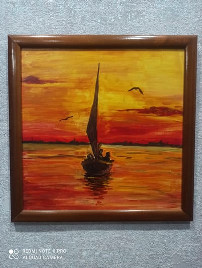 The image displays a framed painting depicting a sunset over a body of water with a sailboat. The sky is a vibrant gradient of yellow, orange, and red, with two birds flying in the distance. A silhouetted shoreline with indistinct structures is visible on the horizon. The water reflects the fiery colors of the sunset. A small sailboat with a dark sail and several silhouetted figures aboard is positioned in the foreground, slightly to the right of center. The painting is framed by a simple wooden frame. The background of the image is a textured grey wall. No information about the city of Kugesi, Russia is present in the image itself.