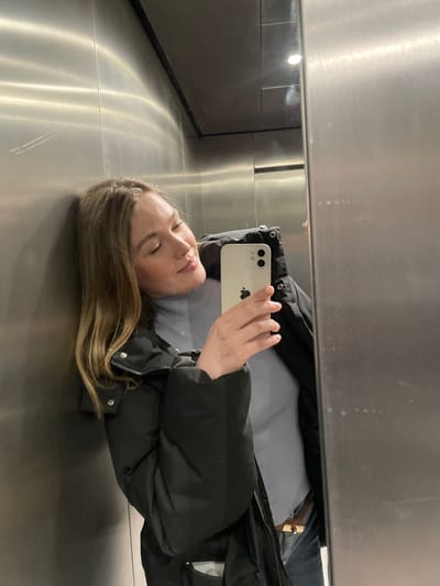A young woman, lying on a reflective surface, possibly metal or glass, takes a selfie with a white iPhone. She is wearing a light gray shirt under a black puffer jacket. Her eyes are closed, and she appears relaxed. The reflection captures part of the surrounding environment, including what may be an elevator interior. The lighting is bright and somewhat diffuse, creating a soft glow on the surfaces.
