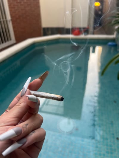 A hand is holding a lit cigarette or marijuana joint against the backdrop of a tiled pool. The hand has long, painted fingernails. Smoke is rising from the lit end of the cigarette/joint. The pool is rectangular and filled with water. The location of the scene cannot be determined with the details provided in the image.