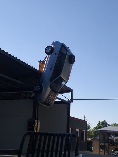 A silver car is displayed upside down, seemingly crashed onto the sloped roof of a building. The car is angled downwards, with its wheels facing the sky. It appears to be a publicity stunt or decorative element for a car wash. The building below has a gray exterior with some red trim and a dark metal roof. A black metal bench is visible in the foreground. In the background, more buildings and trees can be seen under a clear blue sky. The sun is high in the sky, suggesting it is daytime. Text on the car reads "CAR WASH UNTILA" and "Preț - Timp - Calitate" which translates to "Price - Time - Quality". The overall impression is one of a quirky and attention-grabbing advertisement.