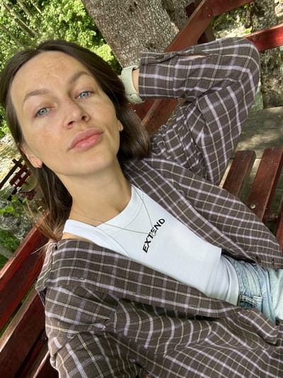 A woman with fair skin, blue eyes, and brown hair is captured from the chest up, sitting on a wooden bench outdoors. She is wearing a brown and purple plaid shirt over a white ribbed tank top with the word "EXTEND" printed in black. She has a delicate silver necklace and small hoop earrings. Her left arm is bent behind her head, and her right arm is resting on the bench. The background consists of a large tree trunk and lush green foliage, suggesting a natural or park-like setting. The lighting indicates it is daytime, with sunlight dappling through the leaves. The image appears to be a selfie, taken at a slightly low angle, with the woman looking directly at the camera. The location is identified as Bare Kraljske, Montenegro.