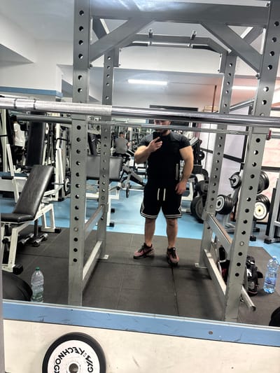 A man wearing a black t-shirt and black shorts with white trim stands in a gym, holding a phone in his left hand, seemingly taking a picture of himself in a mirror. The gym contains weightlifting equipment including a power rack, barbell, weight plates, benches, and other exercise machines. A water bottle is on the floor next to some equipment. The location is a gym in Laureana di Borrello, Italy.
