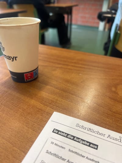 The image shows a desk with a coffee cup from "mayr" brand and a document with the title "Schriftlicher Ausd". The document also has "So sieht die Aufgabe aus" and "30 Minuten Schriftlicher Ausdruck" written on it. The desk is made of wood. The background shows a blurry view of what appears to be a room with chairs and possibly a window. The picture was taken in an unspecified indoor location; the image provides no information about the city of Bayreuth, Germany.