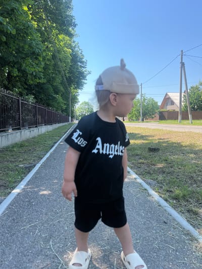 A young child, likely a toddler, stands on a paved path next to a grassy area. The child is wearing a black t-shirt with white lettering that reads "Los Angeles", black shorts, a beige cap, and white slip-on shoes. The child is facing to the right and looking away from the camera. The path is bordered by a low concrete curb. To the left of the path is a dark metal fence running alongside a row of green trees. To the right of the path is a dry, grassy field with some scattered debris. In the background, there are more trees, a utility pole with wires, and a house with a pitched roof. The sky is clear and blue. This scene is situated in the city of Blizhniy Khutor, Moldova.