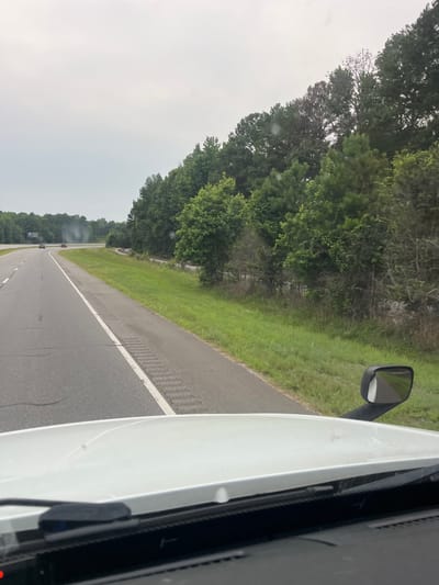 This is a driver's perspective image taken from a white semi-truck, driving on a two-lane highway. The highway is in the foreground, with the truck's hood and windshield wipers visible at the bottom of the frame. The road curves to the right, with a grassy shoulder and a dense line of trees bordering the right side of the highway. In the distance, a red car and a dark-colored car are visible on the highway. The sky is overcast. The location is the city of Arcade, United States of America.