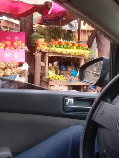 This image captures a street-side market scene, likely in Aketu-Oja, Nigeria, as viewed from inside a vehicle. The primary subjects are the various fruits and vegetables displayed for sale. Piles of red apples, possibly Pink Lady brand given the visible pink boxes with white lettering, are prominent on the left. Beside them, a cluster of pineapples rests on what appears to be a wooden stall.
Further into the scene, a diverse array of vegetables is arranged on a wooden table. This includes bundles of leafy greens, stacked cucumbers, and rows of carrots. Colorful bell peppers—red, yellow, green, and orange—are neatly placed atop a yellow fringed cloth. Some lemons and a bottle of cleaning product are also visible on a lower shelf.
A person, mostly obscured, is standing behind the produce stall. They are wearing a brown garment and their presence suggests they are the vendor. The setting is an outdoor market, indicated by the makeshift stalls and colorful tarps overhead, which provide shade. The lighting suggests it is daytime, with no specific indicators of weather conditions. The overall mood is that of a bustling local marketplace.
The image also shows parts of the car's interior, including the steering wheel, dashboard, and a side mirror reflecting a portion of the market. The visible text on the steering wheel includes "SRS AIRBAG." The pink boxes also bear the "Pink Lady" logo.