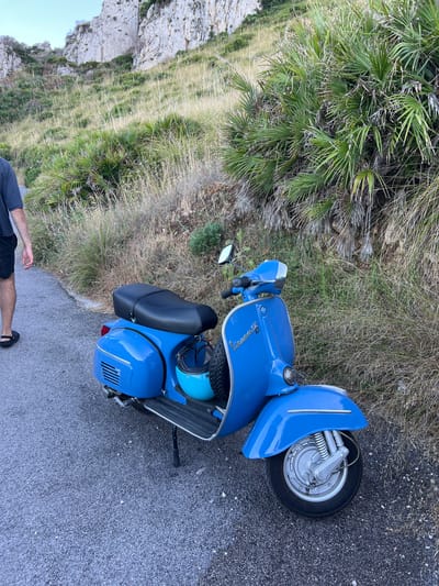 A blue Vespa scooter is parked on a paved road in Sperone, Italy. The scooter has a black seat and a light blue helmet is hanging from the back. The scooter is on a road that is next to a grassy hill with some palm trees. In the background, there are rocky cliffs. A person's legs are visible on the left side of the image, wearing black shorts and black sandals. The person is walking away from the scooter.