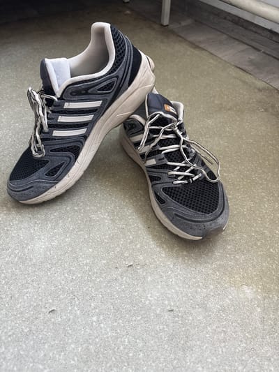 A pair of dark gray and white athletic shoes with white laces are placed on a light gray concrete floor. The shoes are positioned at an angle, with the left shoe slightly in front of the right. The floor appears to be outdoors, possibly a patio or sidewalk, with some dirt and debris present. The background shows a blurred area of wooden planks and a white structure, possibly furniture or a fence. The lighting suggests it is daytime. The shoes are the primary focus of the image, and their condition indicates they have been used. There is no visible indication of the location being within Урыцкі сельскі Савет, Belarus.