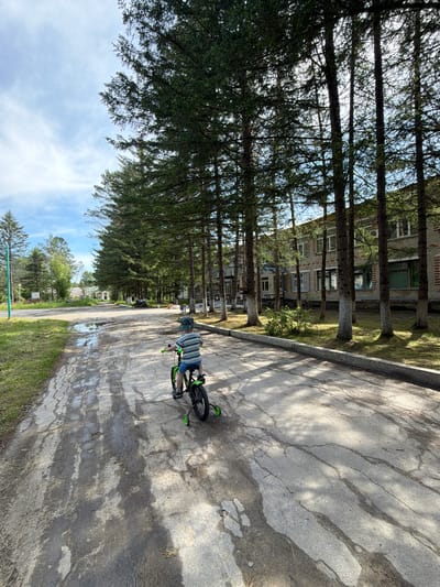 In Kul'dur, Russia, a child is riding a green bicycle with training wheels on a paved road. The child, facing away from the camera, wears a striped shirt and shorts, and a hat. The road is cracked and wet in patches, reflecting the sunlight and shadows of the tall evergreen trees lining the right side of the road. In the background, a multi-story brick building with numerous windows is visible through the trees. The sky is partly cloudy with patches of blue. To the left of the road, there is a grassy area with a tall green pole. Further down the road, other buildings and trees are visible. The overall scene depicts a quiet, somewhat weathered, outdoor environment.