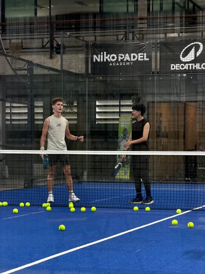 The image depicts an indoor padel tennis court in Ban Buak Khrok North, Thailand. Two young men are visible on the court, separated by the net. The man on the left, wearing a light grey sleeveless shirt and dark shorts, is holding a blue padel racket and appears to be mid-conversation or instruction. The man on the right, dressed in a black sleeveless top and dark pants, also holds a blue padel racket and is looking towards the left. Several yellow padel balls are scattered across the blue court surface. The background shows the facility's infrastructure, including netting, signage for "Niko Padel Academy" and "Decathlon," and glass walls. The lighting suggests an indoor evening or daytime setting.