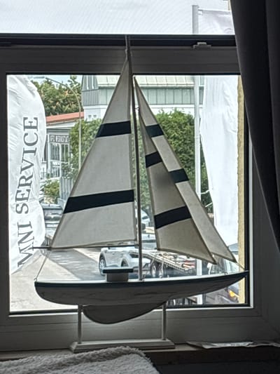 A model sailboat is displayed on a windowsill in front of a window. Outside the window, trees, buildings, and vehicles are visible. To the left of the sailboat, a white banner with the text "MNI SERVICE" is partially visible. The scene appears to be located in Dachau, Germany.