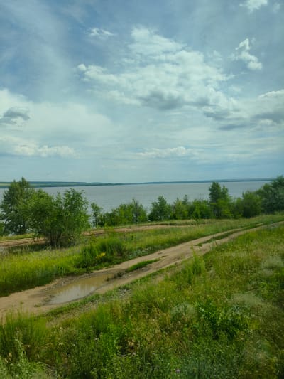 The image shows a wide, calm river under a cloudy sky. The foreground is dominated by a grassy slope with a dirt road winding through it. The road has a large puddle of water on it. There are trees and bushes on the banks of the river. The distant bank of the river is lined with trees and appears to be part of a rural landscape. The scene evokes a sense of natural tranquility and remoteness. The image appears to be a landscape photograph taken in Buzuluk, Russia, possibly near the Samara region, known for its natural beauty and rivers.