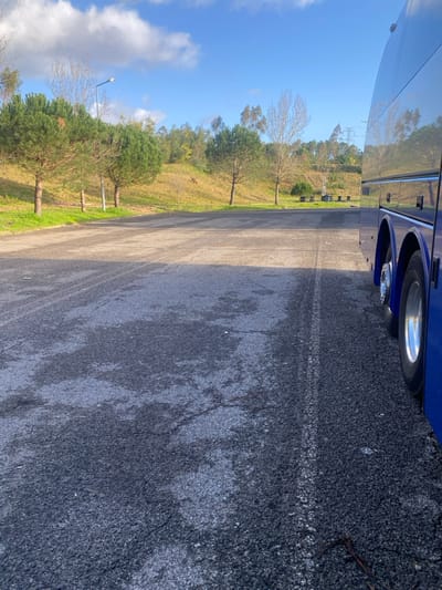 The scene depicts an outdoor environment, specifically a paved parking area or pull-off point in Leiria, Portugal, under clear daylight conditions. The foreground consists of an asphalt surface exhibiting wear, cracks, and irregular lighter patches. A faint white line is visible on the asphalt, running parallel to the right edge of the frame.
On the right side of the image, the polished blue exterior of a large vehicle, identified as a coach or bus, is visible, reflecting the surrounding environment. Its side windows and two chrome-rimmed wheels are discernible. The vehicle is stationary.
The central and left portions of the midground present an open asphalt area, leading to an elevated grassy bank. This bank is covered in dry grass and features numerous trees, including several shorter pine trees with dense green foliage and taller deciduous trees with bare branches. A gray utility pole supporting a street lamp stands vertically on the left side of the bank. Further up the bank, towards the right, a small cluster of light-colored structures resembling benches or picnic tables is present.
The background is dominated by a clear blue sky with scattered white cumulus clouds. Additional trees and utility structures are faintly visible on a distant hillside. No individuals or discernible activities are present in the frame.