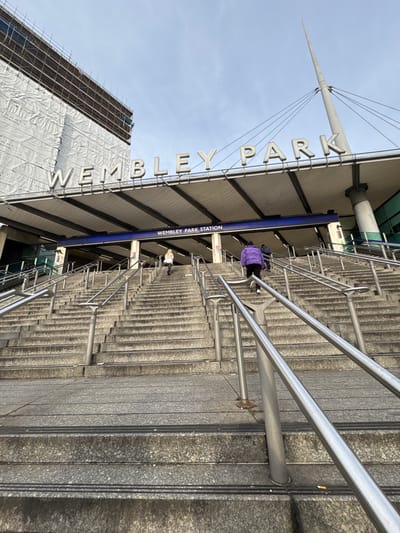 This media file captures a wide-angle view of the exterior entrance to Wembley Park Station in Wembley, United Kingdom, seen from the bottom of a large staircase looking upwards.
The prominent subject is a broad, multi-tiered granite staircase, likely leading to the station's concourse or platforms. Numerous metal railings, both central and along the sides, assist pedestrians. Several people are visible on the stairs; one individual wearing a purple jacket is ascending near the middle, and another person in a white top is further down.
Above the stairs, a large, modern canopy extends, featuring a distinctive pointed mast supported by cables. Spanning the length of the canopy are large, white letters spelling "WEMBLEY PARK". Below this, a darker blue sign clearly displays the text "WEMBLEY PARK STATION".
To the left of the stairs and station entrance, a large building is under construction or renovation, covered extensively in scaffolding and white protective sheeting. The sky above is bright and clear, indicating a daytime setting with fair weather. The scene depicts typical pedestrian activity at a public transport hub.