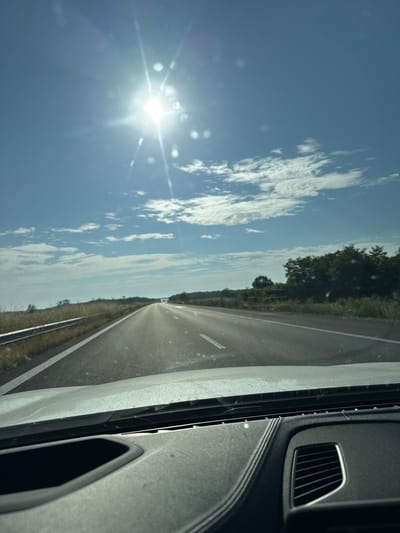 The image displays a forward-facing view from inside a vehicle, looking out onto a highway under bright daylight. The sky is predominantly clear blue with scattered white cumulus clouds, primarily in the upper right quadrant. The sun is intensely bright in the upper-left-center, causing significant lens flare and glare that diffuses light across the upper frame.
The asphalt highway appears straight and extends into the distant horizon, with a noticeable shimmering effect on its surface. White solid lines mark the road's shoulder and potentially separate lanes. To the left, a silver metal guardrail runs parallel to the road, beyond which lies dry, yellowish-brown grass and low vegetation. The right side of the road also features dry grasses, leading to a dense line of green trees in the mid-distance, defining the horizon on that side. No people are visible.
The bottom portion of the image shows the dark gray dashboard of the vehicle. It features distinct textures, an open compartment or speaker grille on the left, and a multi-slatted air vent on the right, consistent with a contemporary car interior. The windshield exhibits minor specks or smudges.
This scene depicts a rural or semi-rural highway environment, characterized by open land, trees, and clear weather. Based on the provided instruction, this highway setting is located within or in the vicinity of Kolarovo, Bulgaria.