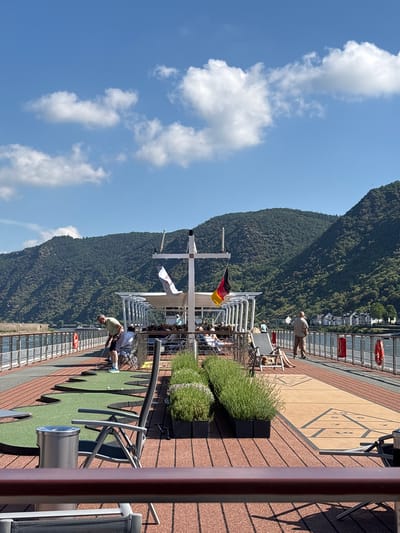 The image depicts a sunny day on a cruise ship deck in Kestert, Germany. The foreground shows a miniature golf putting area with green turf and metal putters. A person in a light green shirt and dark shorts is visible on the putting green. Further back, a shuffleboard court is marked on the deck. Several people are seated in lounge chairs, some under umbrellas, enjoying the scenery. A German flag is prominently displayed on a flagpole.
In the background, a steep, tree-covered mountainside rises dramatically, with houses visible nestled along its base. The Rhine River is implied to be nearby, as suggested by the cruise ship setting and the riverside town. The sky is bright blue with fluffy white clouds, indicating clear and pleasant weather. The sun is casting distinct shadows from the railings and furniture, suggesting it is midday. The overall atmosphere appears relaxed and leisurely, with passengers enjoying the scenic views of the German landscape.