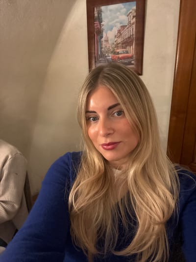 The image depicts an indoor scene in Aix-en-Provence, France. A woman with long, wavy blonde hair, light blue eyes, and reddish-pink lipstick is positioned in the foreground, facing and looking directly at the camera. She wears a dark blue long-sleeved top over a white garment. A partial view of a second individual wearing a light-colored, possibly beige or off-white, ribbed top is visible to the woman's left. On the light-colored wall behind the woman, a rectangular painting in a dark brown frame is centrally displayed. The painting illustrates an urban street scene featuring old colonial-style buildings, an aged red car, and a prominent domed structure, reminiscent of the Capitolio building in Havana, Cuba, under a partially cloudy sky. To the right of the painting, a vertical section of a dark wooden door frame or panel is visible. The environment is illuminated by warm, artificial light.