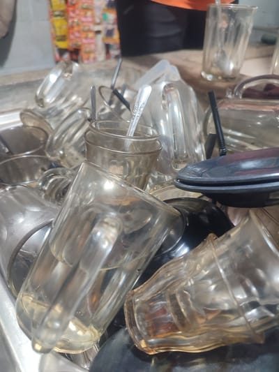 The image depicts a pile of dirty clear glasses and mugs in a sink, likely in a food establishment. The glasses are of various sizes and shapes, some appearing to have residual liquid and residue. In the background, there are shelves stocked with colorful packaged goods, suggesting the location is a shop or warung. The overall scene is unhygienic, with the unwashed dishes creating a cluttered and messy appearance. The lighting is dim, and the focus is primarily on the glasses in the foreground. The specific location in Muliengmanyang, Indonesia, is indicated by the context provided, but the image itself does not offer any direct visual cues to confirm this.