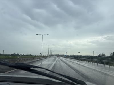 The image captures a view from inside a vehicle driving on a wet, multi-lane highway under a heavily overcast, cloudy sky. The road surface is reflective and shows tire tracks, indicating recent rain. Streetlights line the highway at regular intervals, and a concrete barrier with some orange markers separates the lanes from the roadside. The roadside features green vegetation and a line of trees in the distance. The windshield wipers are visible in the foreground, suggesting the rain is ongoing or has just ceased. There are no people or other vehicles clearly visible in the frame. The overall impression is one of a gloomy, wet day on a highway. The specific location in the city of ქალაქი ლანჩხუთი, Georgia, cannot be determined from the visual information present in the image.