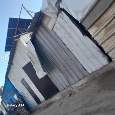 The scene depicts a structure in the urban Al-Mawasi, Unknown. The structure is predominantly constructed of corrugated metal. A solar panel array is mounted on the roof. The photograph has been taken from a low angle, giving a distorted perspective. The structure is partly obscured by tarpaulin. The ground visible is made of concrete. The photo has text overlay: "Galaxy A14".