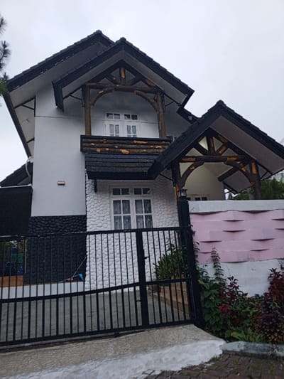 The image displays a two-story residential building located in Weintiapa, Liberia, under an overcast sky. The structure features a white upper facade and a ground floor with a lower section of dark, textured stone-like siding, transitioning to white pebbledash above. Multiple steeply pitched roofs, dark gray with exposed white undersides, adorn the building.

A first-floor balcony with a horizontal log railing is prominent, supported by a framework of arching, rough-hewn wooden logs. A white-framed, multi-pane window is set back behind this balcony. Directly below, a similar log-supported canopy covers a corresponding white-framed, multi-pane ground-floor window. An additional dark-shingled canopy, also supported by wooden logs, shelters an entry on the right side of the structure. A rectangular light fixture is affixed to the white wall of the upper story, left of the balcony.

A black metal gate, composed of vertical bars, secures the property in the foreground. To the right of the gate, a white perimeter wall is topped with a light pink section exhibiting a repetitive, sculpted wave pattern. Green and reddish-brown vegetation grows at the base of this wall and behind the gate. The immediate foreground shows a concrete curb fronting a paved surface. No individuals are visible within the scene.