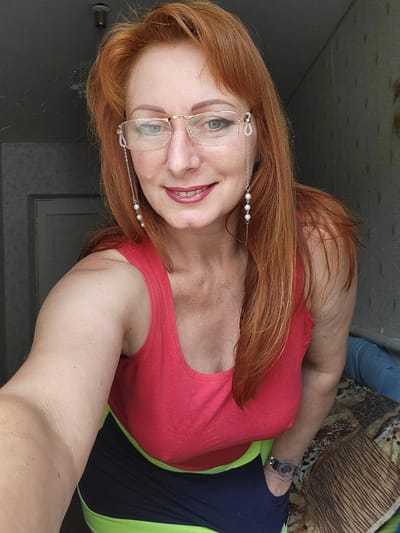 A woman with long, wavy, reddish-orange hair and fair skin is looking into the camera and smiling. She is wearing glasses with decorative pearl and chain embellishments on the sides. Her lips are painted with a bold, berry-colored lipstick. She is wearing a bright pink tank top with a black and neon green sports bra underneath. A watch with a silver band and a purple face is visible on her left wrist. She is positioned in front of a grey-patterned wall. To her right, there is a bed with a patterned comforter. A portion of a dark wooden door is visible to her left. The scene appears to be indoors, likely in a residential setting within Zolotonosha, Ukraine.
