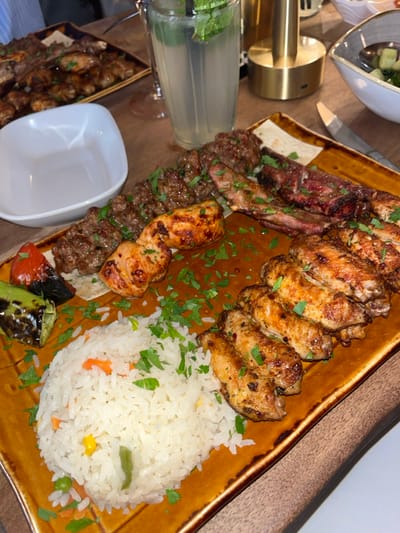 This image captures a dining table laden with food within an indoor restaurant setting, consistent with establishments found in Stevenage, United Kingdom. The central focus is a large, rectangular, amber-brown ceramic platter presenting a mixed grilled meal. This platter contains two skewers of what appears to be minced meat kebabs, several pieces of grilled chicken (including multiple chicken wings), and grilled lamb cutlets or ribs. A generous portion of white rice mixed with diced carrots, peas, corn, and green beans is positioned on the lower-left of the platter, alongside grilled red and green bell pepper pieces. All grilled items and the rice are garnished with chopped fresh parsley. A reddish-brown sauce or marinade is visible beneath some of the meats.
To the upper left, a second, darker rectangular platter holds additional grilled chicken wings and other meat pieces. A white, empty, rectangular bowl with rounded corners is placed mid-left. A tall, fluted glass containing a light-colored beverage, garnished with fresh mint leaves and equipped with a straw, stands in the upper center. A brass-colored cylindrical table lamp is visible on the upper right. Partially visible on the far right are a small grey bowl containing a green salad (likely diced cucumber) and a knife. The table surface beneath the dishes appears to be made of wood or a wood-effect material. No individuals are visible in the scene. The specific restaurant establishment within Stevenage cannot be identified from the visual content of the image.